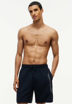 Young man with short hair wearing black Tommy Hilfiger swim shorts standing against a plain light background.