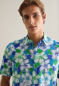 Camisa de manga corta hecha de tela con un patrón floral en azul, verde y blanco, con cierre de botones y cuello puntiagudo.