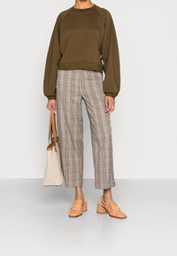 Olivově zelená mikina s raglánovými rukávy, béžové široké kalhoty s kostkovaným vzorem, světle hnědé boty na blokovém podpatku a světlá taška přes rameno.