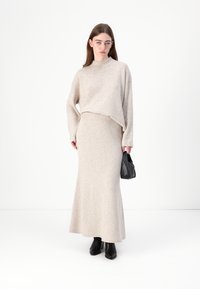 Beige strikkegenser med avslappet passform og høy hals, sammen med et matchende langt skjørt. Modellen holder en liten svart veske, og har på seg svarte støvler.