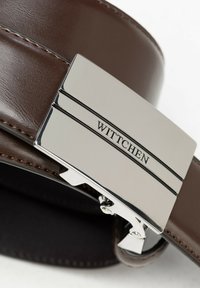 Brązowy skórzany pasek z błyszczącą srebrną metalową klamrą wygrawerowaną napisem "WITTCHEN", z eleganckim, nowoczesnym wzornictwem zapięcia i detalami zszycia.
