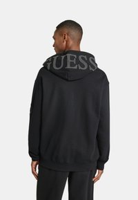 Felpa nera con cappuccio e grande logo "GUESS" grigio sul cappuccio, con una vestibilità comoda e polsini e orlo a coste.
