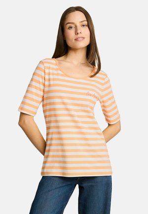 Młoda kobieta z prostymi brązowymi włosami nosi koszulkę na krótki rękaw w paski brzoskwiniowo-białe oraz niebieskie dżinsy, stoi ze złożonymi za plecami rękami.