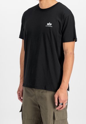 Mężczyzna ubrany w czarną koszulkę Alpha Industries oraz oliwkowo-zielone spodnie typu cargo, stojący na białym tle.