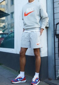 Grå sweatshirt med orange Nike-logotyp, kombinerad med ljusgrå strukturerade shorts och röda, blå och vita Nike sportskor.