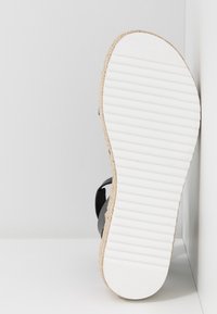 Sandale noire avec une semelle intermédiaire en jute tressé, dotée d'une semelle extérieure en caoutchouc blanc à rainures et d'un design plat. Positionnée en angle.