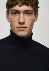Schwarzer Rollkragenpullover mit strukturiertem Strickdesign, figurbetontem Schnitt und hohem Kragen. Das Model hat lockige Haare und einen neutralen Gesichtsausdruck.