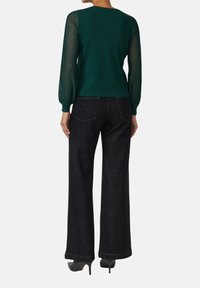 Frau in einem dunkelgrünen Langarmoberteil mit transparenten gestreiften Ärmeln und hoch taillierten, weit geschnittenen schwarzen Jeans mit Absätzen.