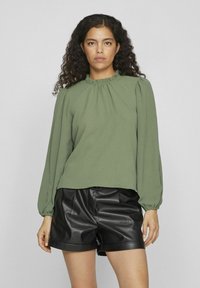 Olijfgroene top met lange mouwen, een verzamelde halslijn en textuurstof, gecombineerd met zwarte faux leren shorts met hoge taille.