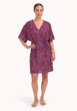 Frau steht barfuß und trägt ein knieumspieltes Kleid mit V-Ausschnitt und gebundener Taille in Lila und Pink mit Muster, lächelt und hat eine Hand in der Hüfte.