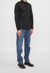 Homme portant une chemise noire à manches longues boutonnée, un jean bleu et des chaussures noires à bout carré, debout devant un fond blanc uni.