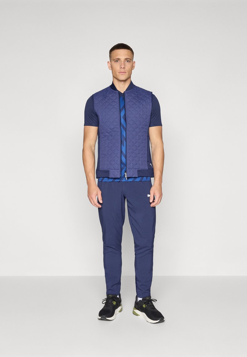 Chaleco acolchado azul marino con un cuello alto sobre una camisa de manga corta azul marino que presenta un patrón geométrico azul, combinado con pantalones azul marino y zapatillas negras.