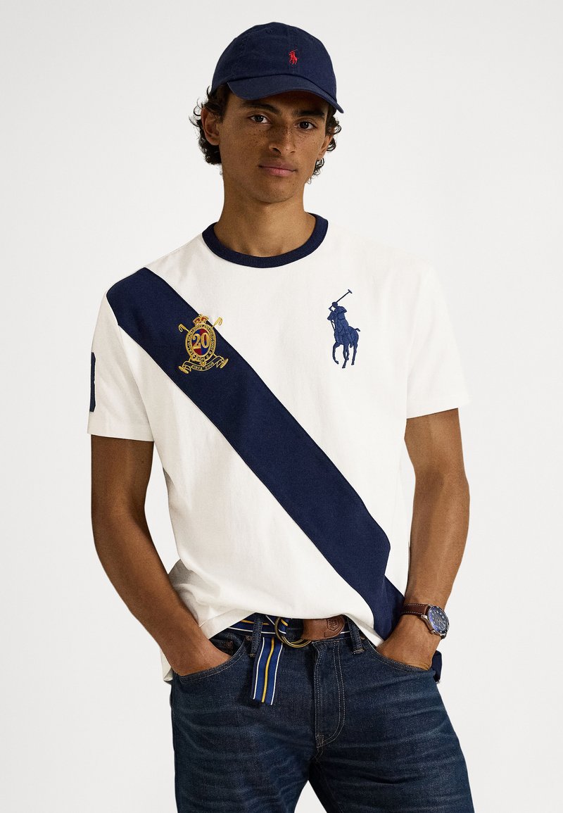 Λευκό βαμβακερό μπλουζάκι με ναυτικό διαγώνιο ρίγα, με κεντημένο έμβλημα και παίκτη του πόλο. Φορεμένο με σκούρο τζιν και ναυτικό καπέλο.