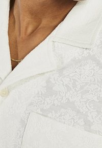 Witte shirt met textuur en een bloemmotief, voorzien van een kraag en knoopsluiting. De stof lijkt zacht en lichtgewicht.