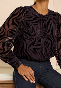 Blusa nera trasparente con un motivo a zebra in velluto, dotata di maniche lunghe e spalle arricciate. Indossata sopra un paio di jeans in denim blu scuro.