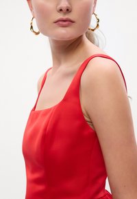 Červené saténové šaty so štvorcovým výstrihom a tenkými ramienkami. Majú hladkú textúru a priliehajúci korzet, ktorý ukazuje jemný sklon. Zlaté kruhové náušnice.