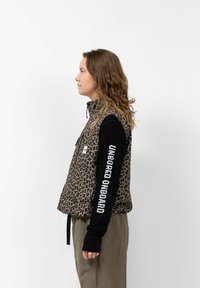 Luipaardprint vest met een hoge kraag, zwarte mouwen met witte tekst. Het materiaal lijkt zacht, met een ontspannen pasvorm en een voorkantzak.