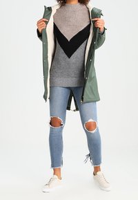 Langer grüner Parka mit Fellfutter, grau-schwarzer Rippstrickpullover mit V-Muster, ausgewaschene blaue Jeans und helle Sneakers.