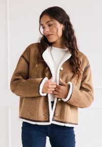 Giacca in suede marrone con un colletto e polsini in ecopelliccia bianca, bottoni frontali e dettagli in piping marrone. Vestibilità casual, lunghezza cropped.