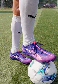 Lila och rosa fotbollsskor med texturerad överdel, snören och en framträdande logotyp, placerade på en flerfärgad fotboll på gräs.