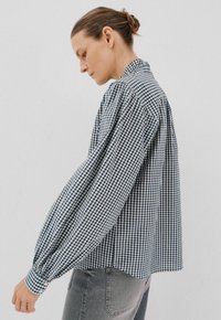 Vrouw in profiel die een zwart-wit geruit overhemd met lange mouwen en een grijze spijkerbroek draagt, met haar in een knot, die omlaag kijkt.