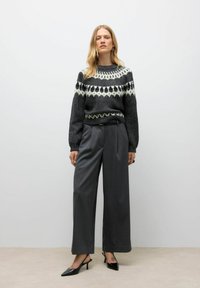 Donna che indossa un maglione a fantasia bianco e nero, pantaloni grigi a gamba larga e décolleté nere a punta, in piedi di fronte a uno sfondo semplice.