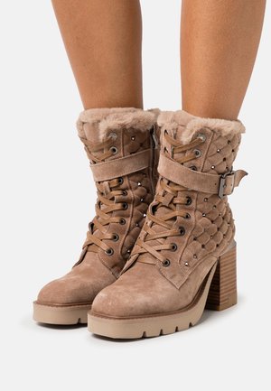 Bottes à talon haut beiges avec des poignets en fausse fourrure, un devant lacé, des accents cloutés et un matériau en daim texturé. Talon carré et semelle épaisse.