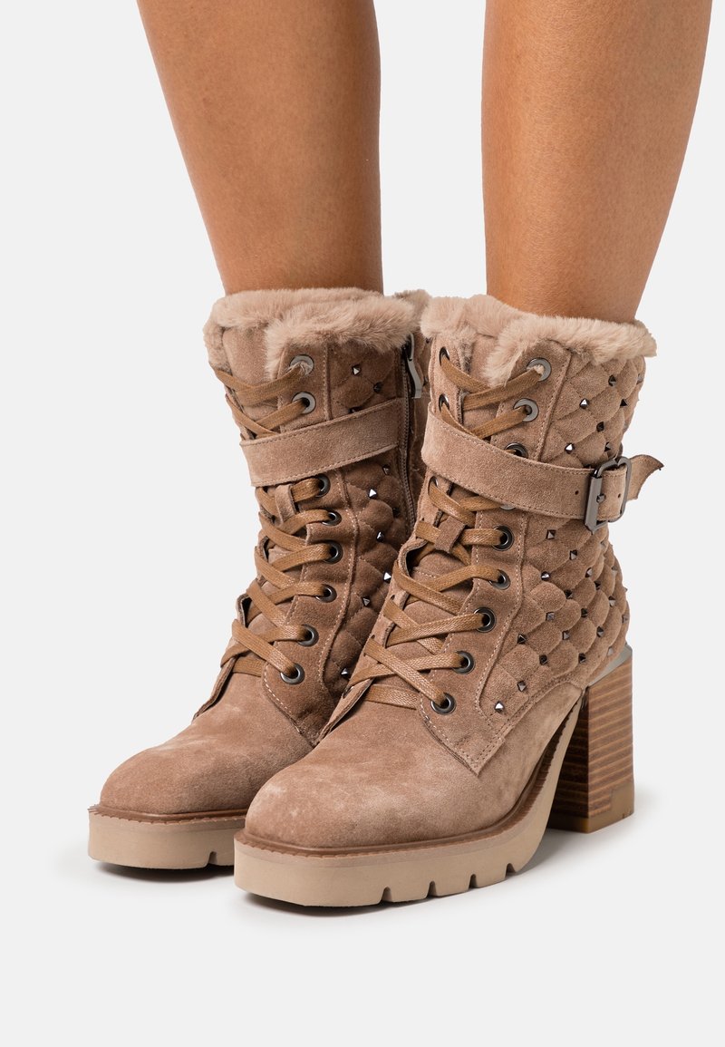 Bottes à talon haut beiges avec des poignets en fausse fourrure, un devant lacé, des accents cloutés et un matériau en daim texturé. Talon carré et semelle épaisse.