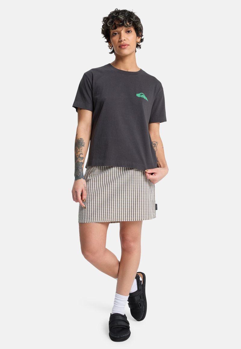 Άτομο που στέκεται με κοντά σγουρά μαλλιά, φοράει μαύρο μπλουζάκι, καρό φούστα, λευκές κάλτσες, μαύρα σανδάλια και εμφανή τατουάζ στα χέρια.