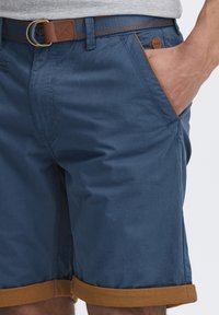 Mørkeblå shorts med en brun oppbrett, sidelommer og en brun læraktig detalj. Belte med en rund metallspenne.