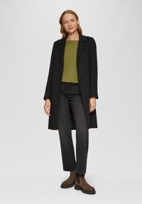 Zwarte wollen jas met een enkele knoopsluiting, gedragen over een groene geribde top en zwarte jeans. Bruine enkellaarzen met een chunky zool maken de look compleet.