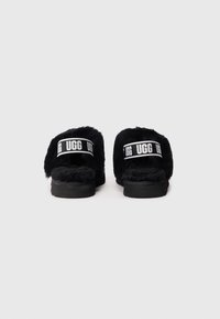 Sorte UGG slide sandaler med fluffy tekstur, bred stropp med logo og en svart gummisåle. Baksidevisning viser åpen hældesign.
