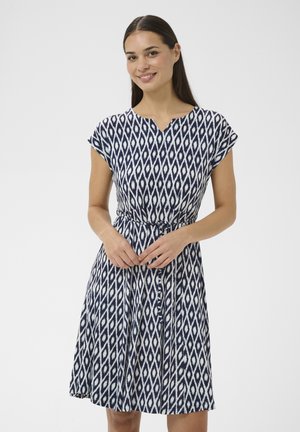 Donna che indossa un vestito a fantasia geometrica blu navy e bianco, lungo fino al ginocchio, con maniche corte, in piedi davanti a uno sfondo bianco uniforme.