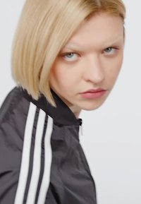 Čierna bunda na zips s bielymi pruhmi na rukávoch, s vysokým golierom a hladkým, lesklým materiálom. Model stojí mierne bokom.