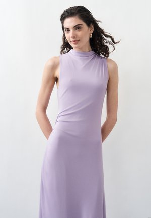 Lokkis juustega naine, kes kannab varrukateta lavendlivärvi kleiti, seisab valge ühtlase tausta ees, käed selja taga.