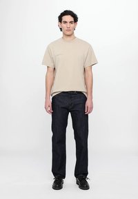 Tricou bej din bumbac cu mâneci scurte și imprimare minimală, asortat cu blugi denim de un albastru închis și pantofi negri cu șireturi. O ținută simplă, lejeră.