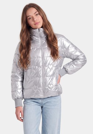 Junge Frau mit langen braunen Haaren, die eine glänzende silberne Steppjacke mit Herzmustern und hellblaue Jeans trägt, steht vor einem einfarbigen Hintergrund.