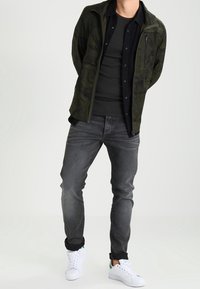 En man som bär en grön kamouflagejacka över en svart skjorta, mörkgrå jeans och vita sneakers, står med händerna bakom ryggen.