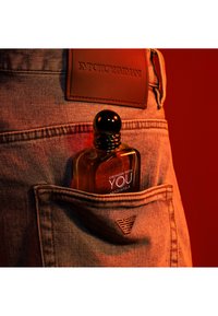 Parfumfles met een ronde zwarte dop die gedeeltelijk zichtbaar is in een denim zak. Het etiket zegt "Stronger With You Absolutely." Rode achtergrond.