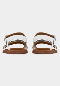 Paire de sandales en cuir blanc avec semelles marron, brides autour de la cheville et boucles argentées, vues de dos sur un fond uni.