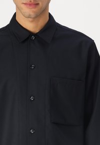 Black button-up shirt made of smooth fabric, featuring a pointed collar, front pocket, and matching black buttons.