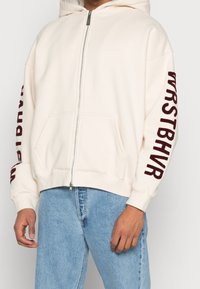 Krémsvěrá mikina na zip s vínově červeným textem na rukávech, přední kapsy, žebrované manžety a kapuce se šňůrkou. Měkká textilie.