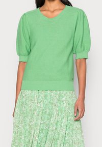 Zelený pletený svetr s krátkými, nafouklými rukávy. Texturovaný, žebrovaný výstřih a lem. Nositý přes odpovídající sukni s květinovým vzorem.
