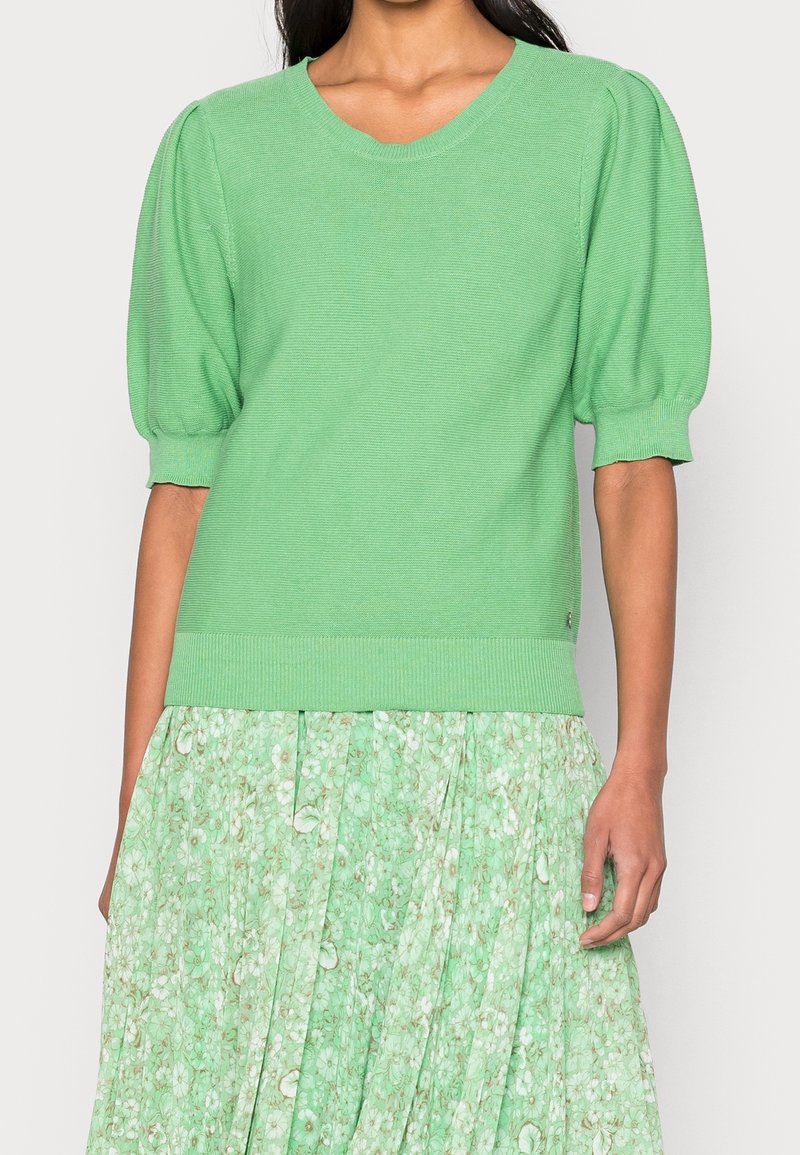 Zelený pletený svetr s krátkými, nafouklými rukávy. Texturovaný, žebrovaný výstřih a lem. Nositý přes odpovídající sukni s květinovým vzorem.