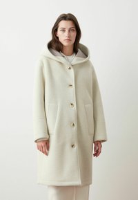Cappotto in misto lana color crema chiaro con cappuccio, grandi bottoni e tasche laterali. Presenta una forma strutturata e una consistenza morbida.