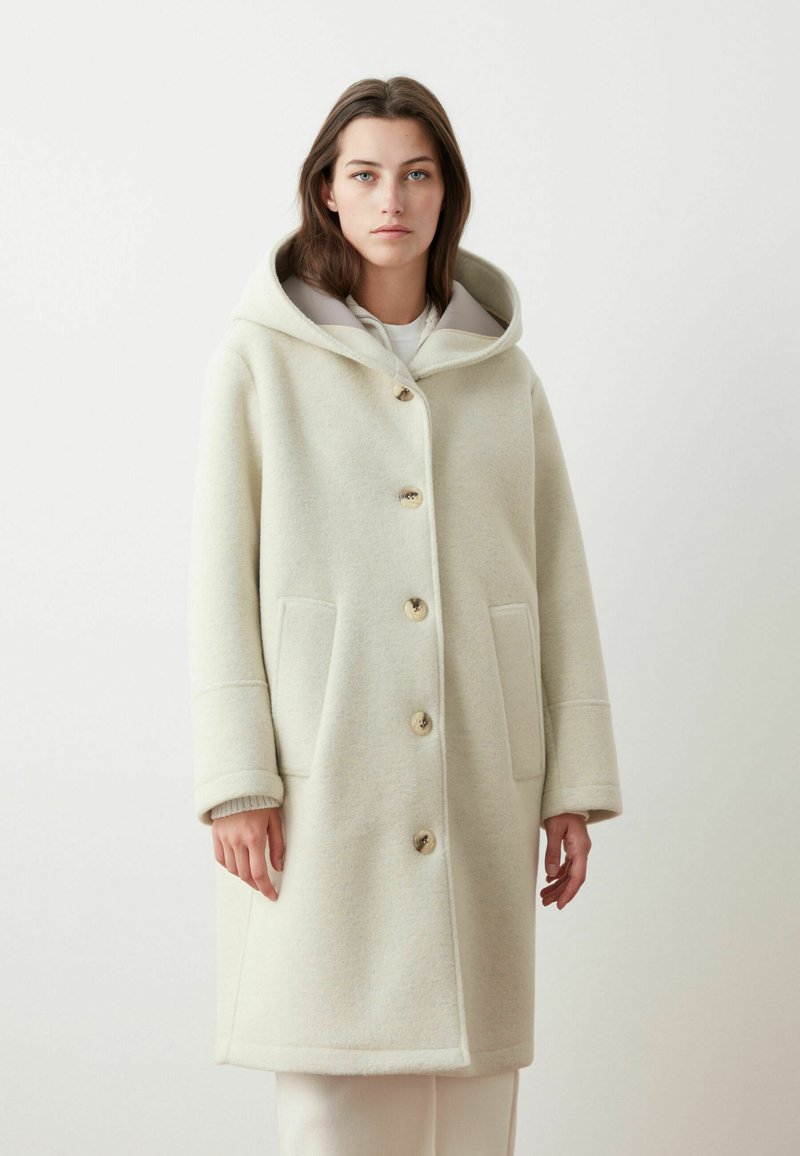 Cappotto in misto lana color crema chiaro con cappuccio, grandi bottoni e tasche laterali. Presenta una forma strutturata e una consistenza morbida.