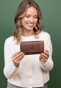 Joven mujer sonriendo con un suéter blanco, sosteniendo una cartera de cuero marrón con ambas manos frente a un fondo verde sólido.