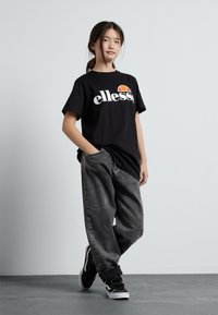 Juoda medvilninė marškinėliai su "ellesse" logotipu ir grafika, derinami su laisviomis pilkos spalvos džinsomis ir juodais sportbačiais su baltais padais.