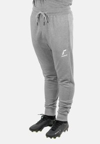 Graue Jogginghose mit elastischem Bund und Kordelzug; verfügt über enganliegende Bündchen und Seitentaschen. Getragen mit schwarzen Stollenschuhen, die mit grünen Stollen akzentuiert sind.