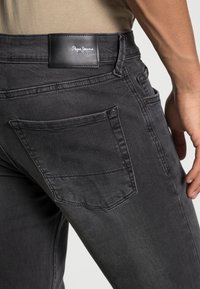 Vaqueros oscuros de corte ajustado, con un parche de cuero con logo, detalles de costura y un bolsillo trasero único con cierre de botón.